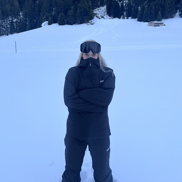 Kvinde med Glaciér skibriller i sort på snedækket piste med skov i baggrunden