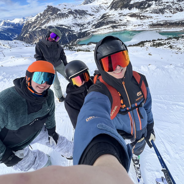 Fire skiløbere med Glaciér skibriller i snedækket bjerglandskab med turkisblå sø i baggrunden