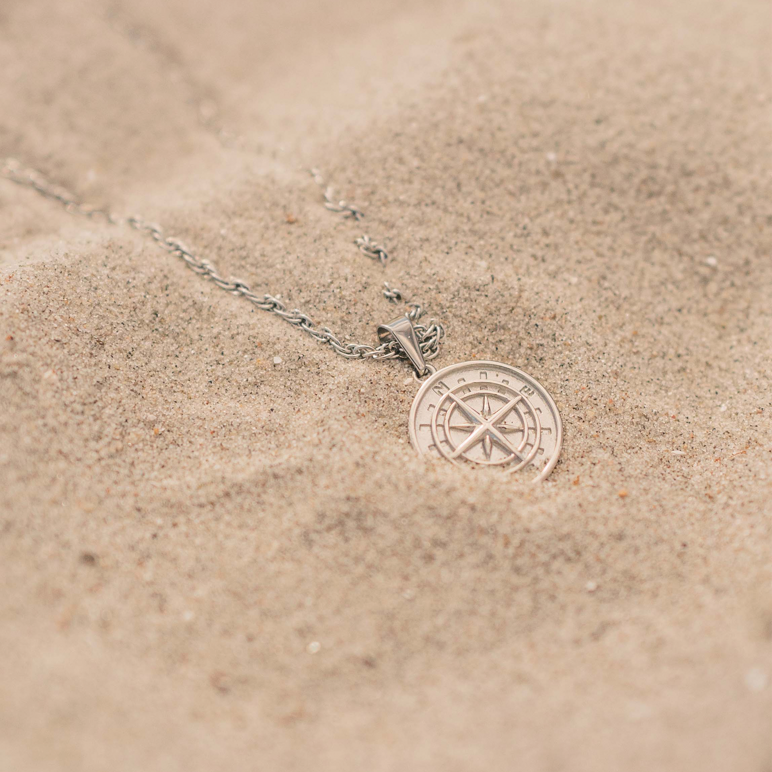 Sølvfarvet halskæde med kompas-vedhæng på sandstrand