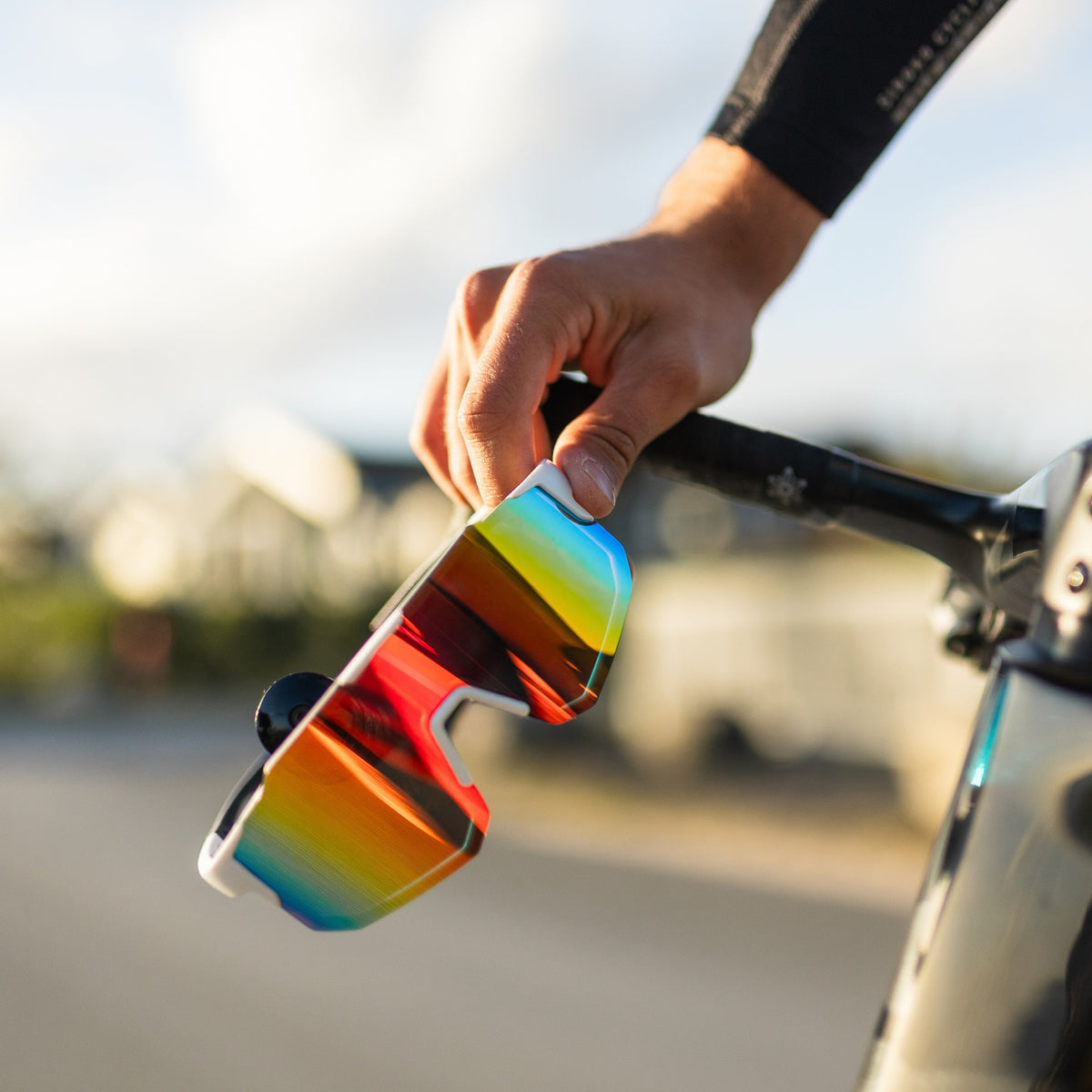 Noire Ruby sportsbriller med tropisk farverig spejllinse holdt i hånden ved cykel
